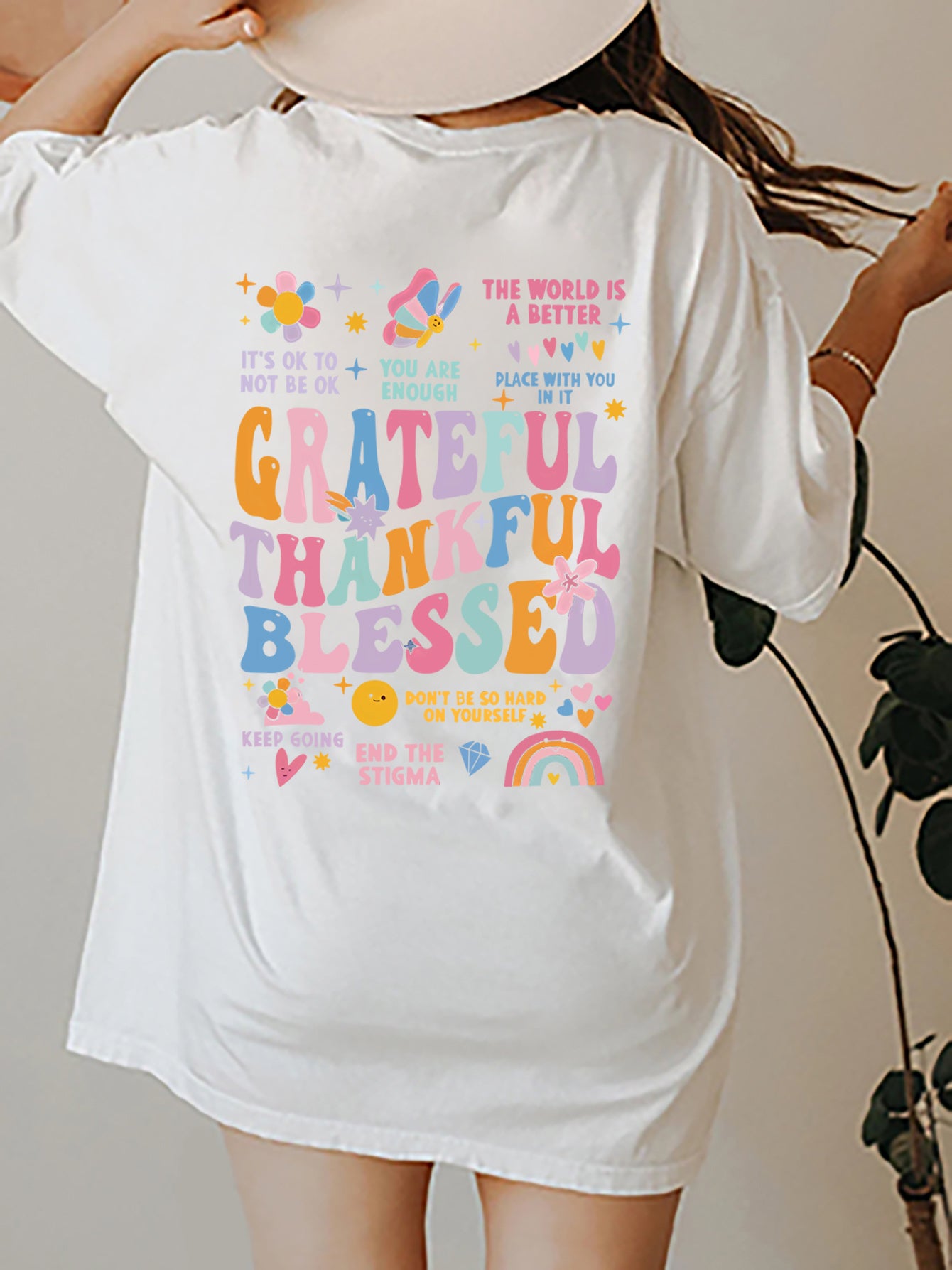 Back view of an oversized white t-shirt with colourful text and graphics reading 'Grateful Thankful Blessed' surrounded by inspirational phrases such as 'You Are Enough', 'It's Ok to Not Be Ok' and 'End the Stigma', along with cheerful design of flowers, butterflies, heats and rainbows.