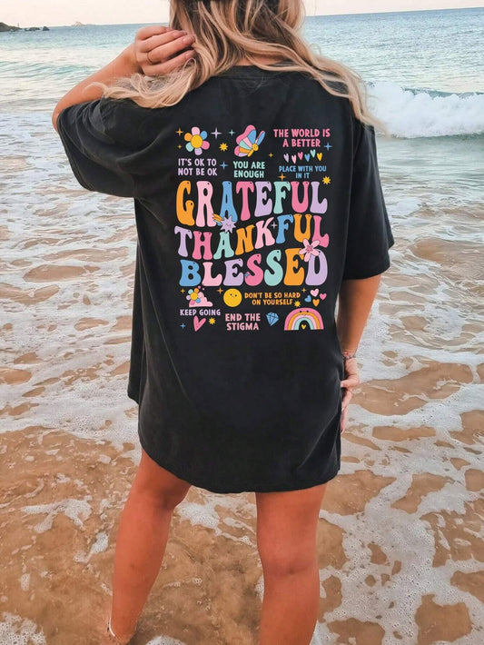 Back view of an oversized black t-shirt with colourful text and graphics reading 'Grateful Thankful Blessed' surrounded by inspirational phrases such as 'You Are Enough', 'It's Ok to Not Be Ok' and 'End the Stigma', along with cheerful design of flowers, butterflies, heats and rainbows.