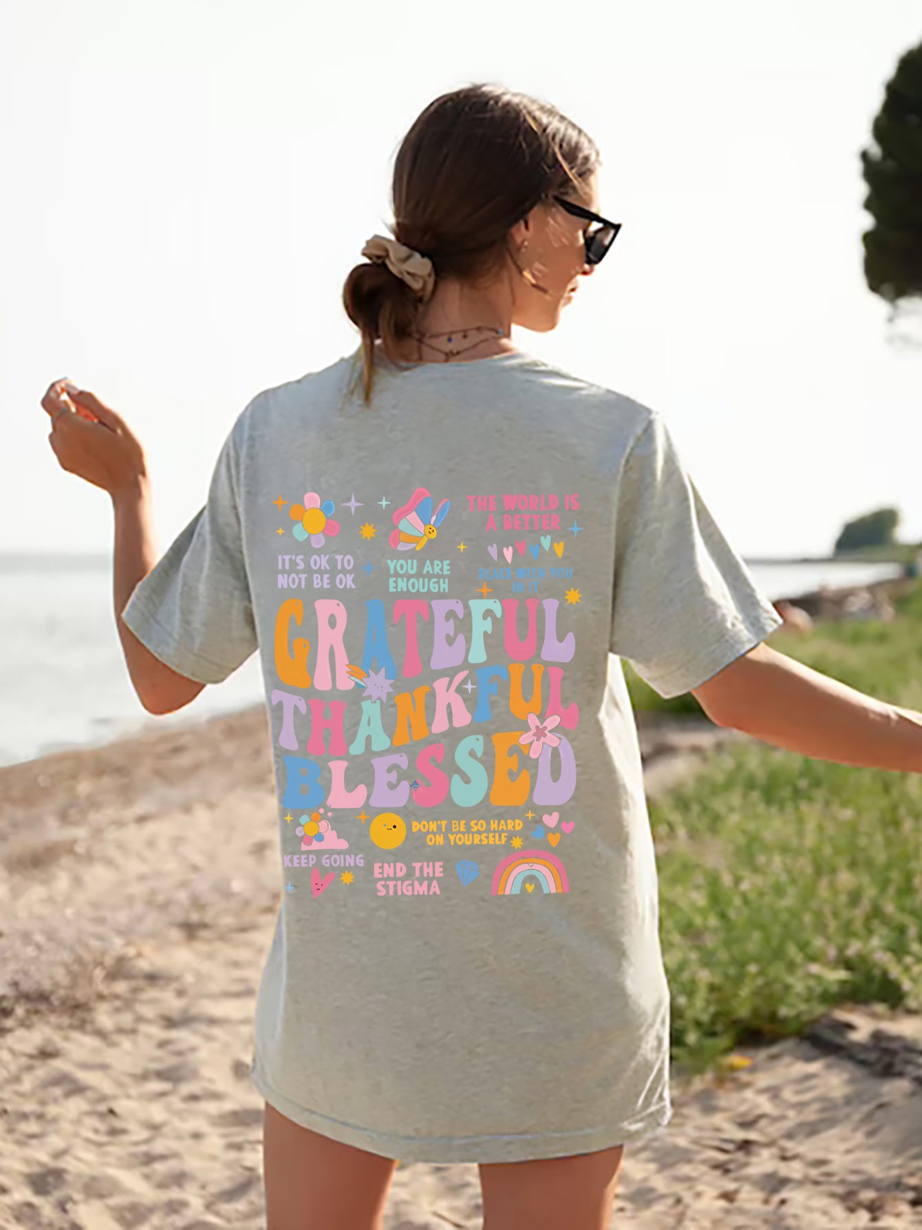 Back view of an oversized light grey t-shirt with colourful text and graphics reading 'Grateful Thankful Blessed' surrounded by inspirational phrases such as 'You Are Enough', 'It's Ok to Not Be Ok' and 'End the Stigma', along with cheerful design of flowers, butterflies, heats and rainbows.